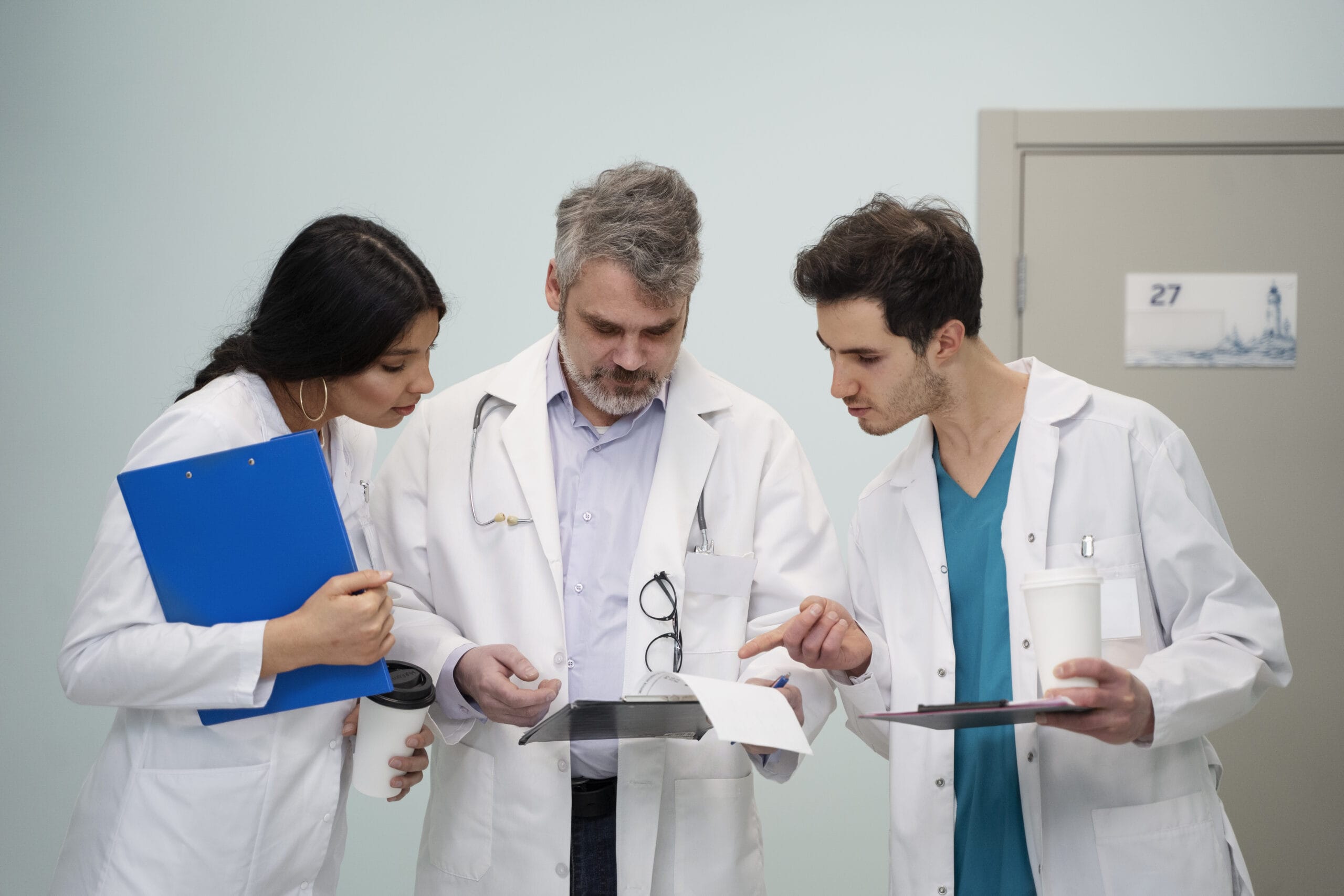 Three healthcare professionals in white lab coats discuss medical documents in a hallway. One holds a clipboard, another reads papers, and the third points to a tablet, highlighting the perks of being a healthcare preceptor through on-the-job clinical teaching advantages.