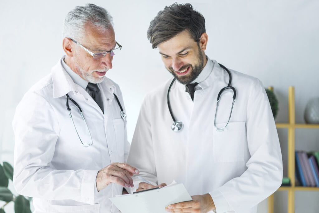 Two doctors in white lab coats are discussing a medical chart together in a brightly lit room, both wearing stethoscopes around their necks. This scene illustrates the benefits of mentorship in medicine, as one doctor takes on a medical mentor role to help develop the other's medical skills.
