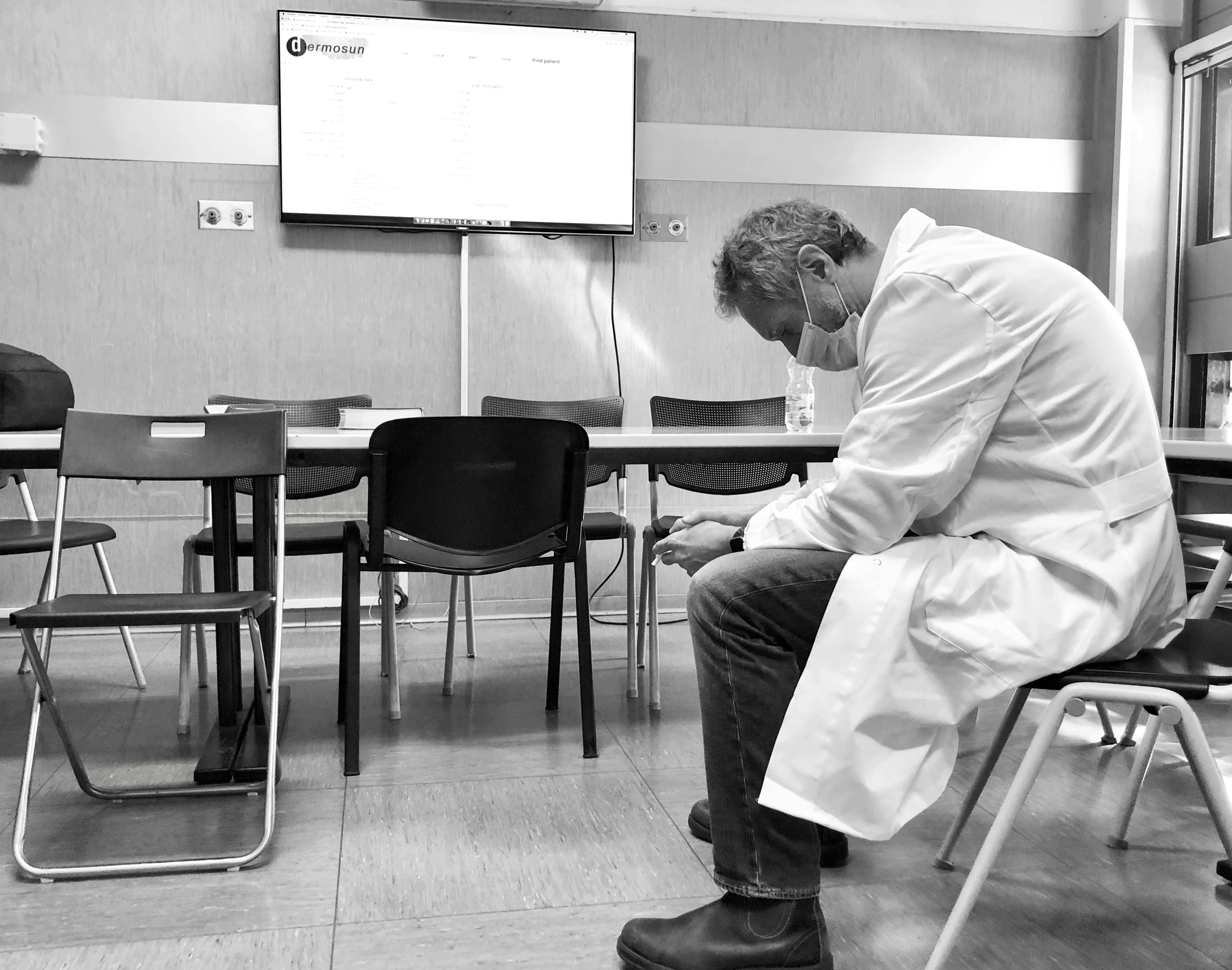 A person in a lab coat and mask sits hunched over on a chair in an empty room, highlighting physician burnout, with a TV screen displaying information in the background.